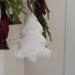 A fluffy white Christmas tree ornament hanging from green garland with a burgundy ribbon and a small brass bell nearby.