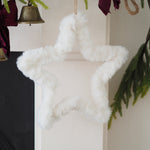 A fluffy white faux-fur star-shaped Christmas ornament hanging against an off-white wall, with holiday decor including greenery, a bell, and burgundy fabric visible around it.