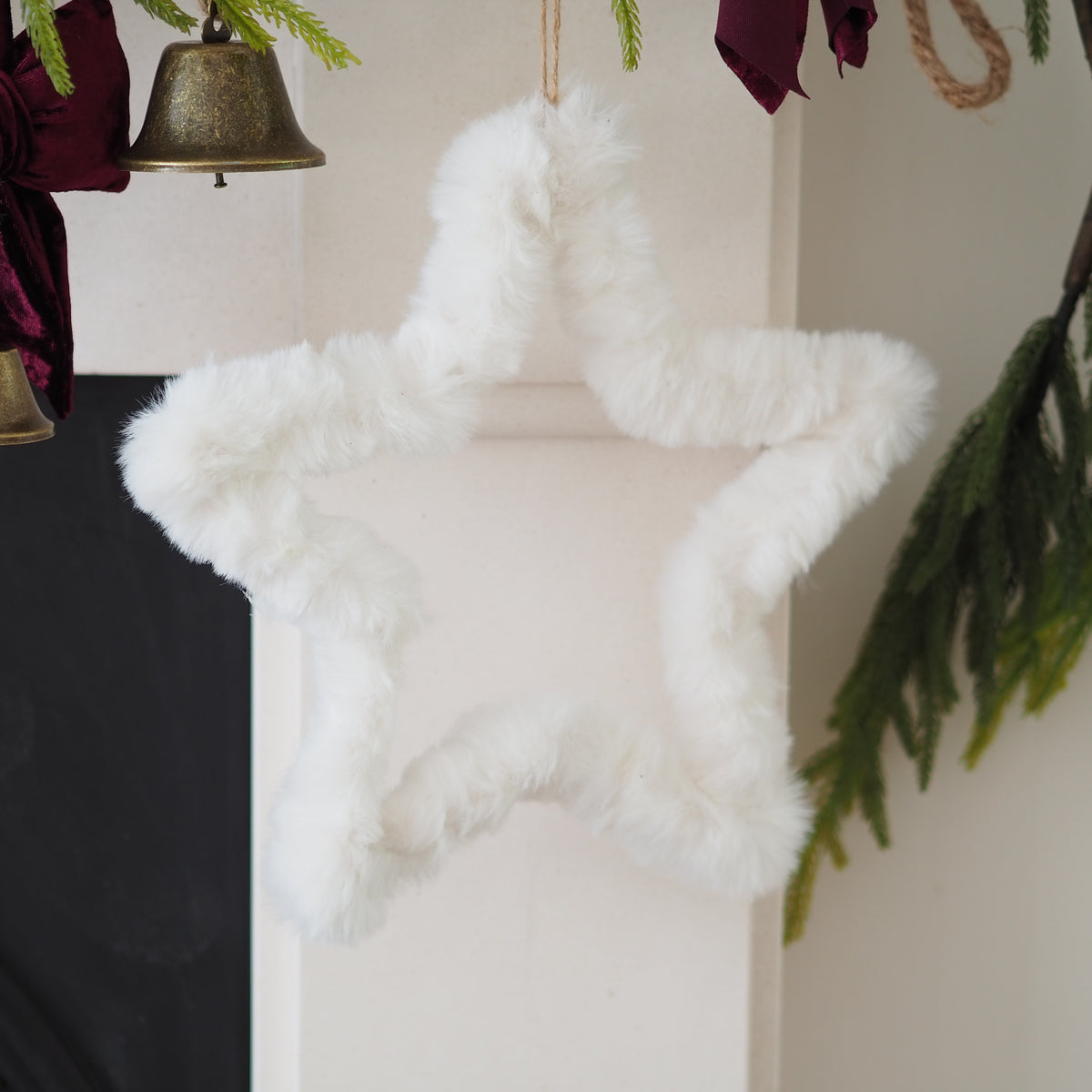 A fluffy white faux-fur star-shaped Christmas ornament hanging against an off-white wall, with holiday decor including greenery, a bell, and burgundy fabric visible around it.