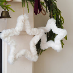 Two fluffy white faux-fur star-shaped ornaments hanging from a green garland with a small brass bell and burgundy ribbon in the background.