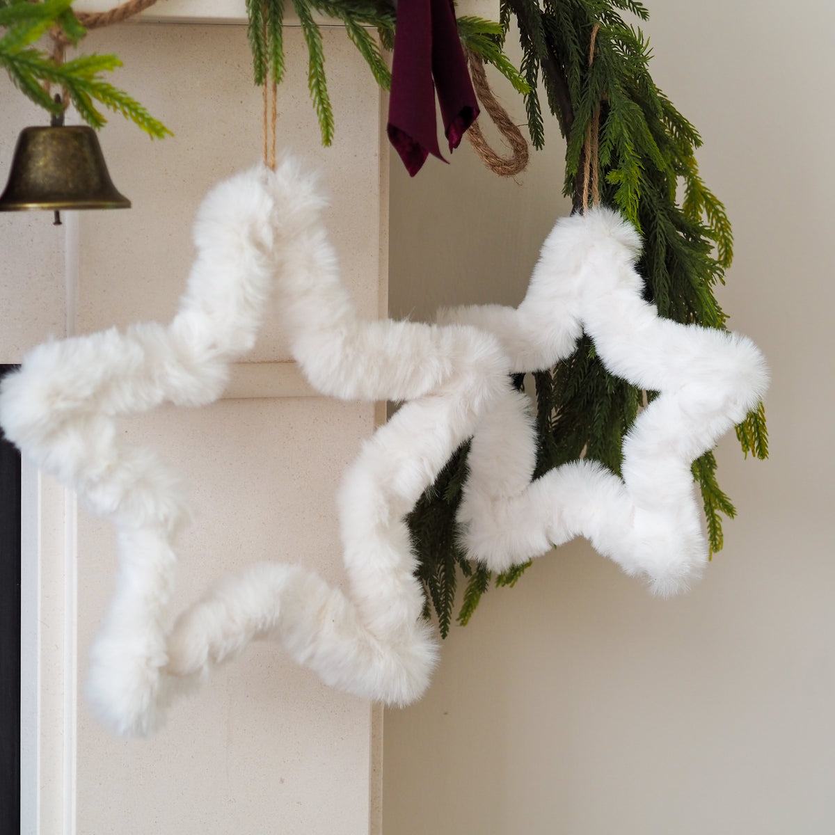 Two fluffy white faux-fur star-shaped ornaments hanging from a green garland with a small brass bell and burgundy ribbon in the background.