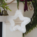 A fluffy white star-shaped Christmas ornament hanging from a rope on a mantel, surrounded by green pine branches, a dark burgundy ribbon, and a small brass bell.