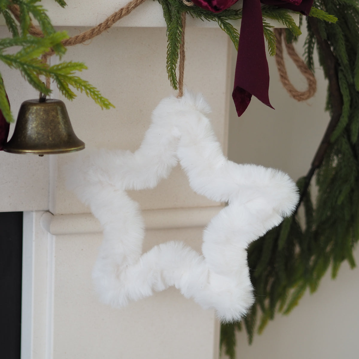 A fluffy white star-shaped Christmas ornament hanging from a rope on a mantel, surrounded by green pine branches, a dark burgundy ribbon, and a small brass bell.