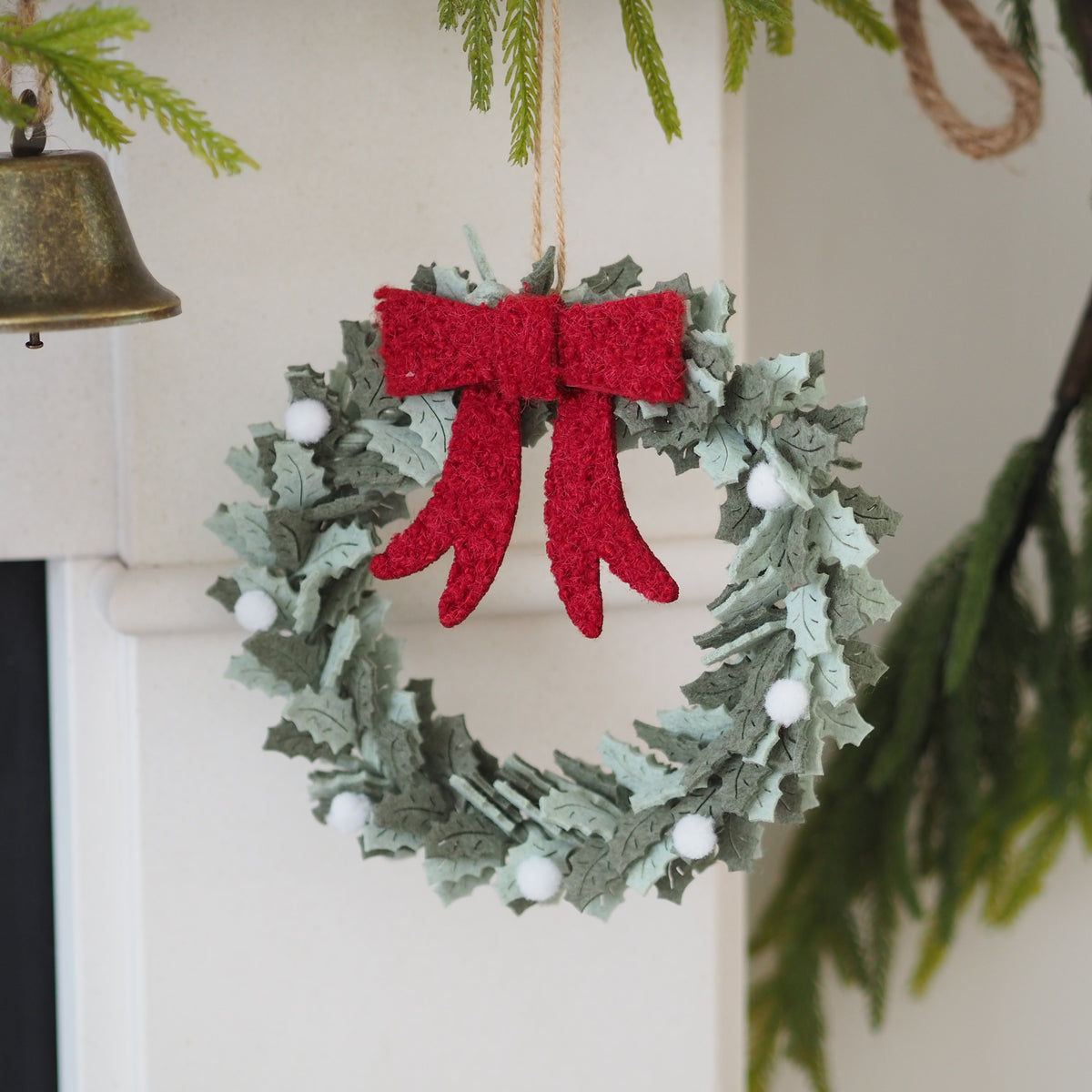 A felt holiday wreath ornament hanging in front of a white fireplace mantel. The wreath is made of sage green and gray-green felted pine branches with a prominent textured red felt bow on top and small white felted pom-poms scattered around.