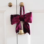 A deep burgundy velvet bow decoration hanging from a brass doorknob on a white paneled door, with a small gold bell dangling below.