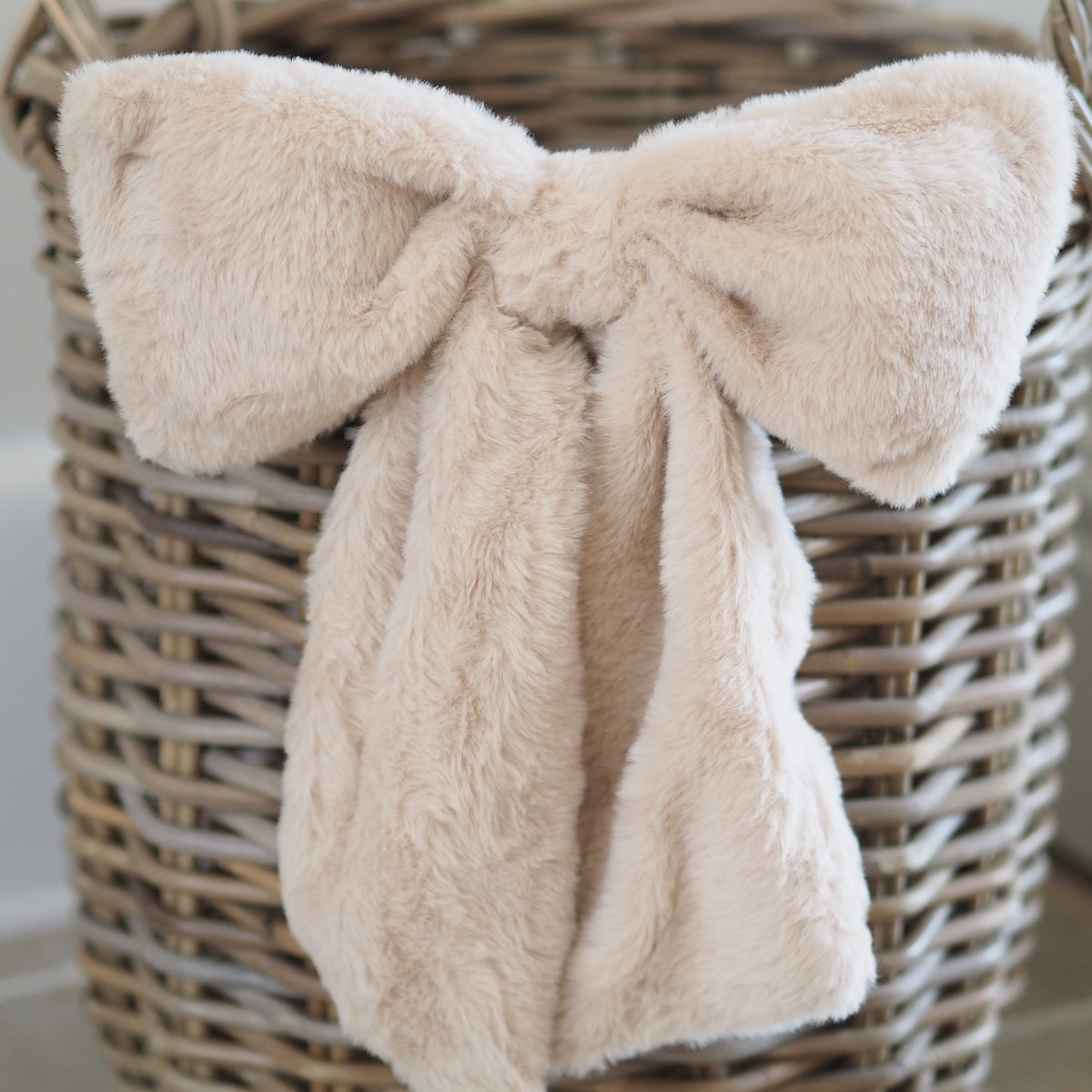 A soft, plush faux-fur throw blanket in a light beige or cream color, styled in a large bow inside a woven wicker basket.