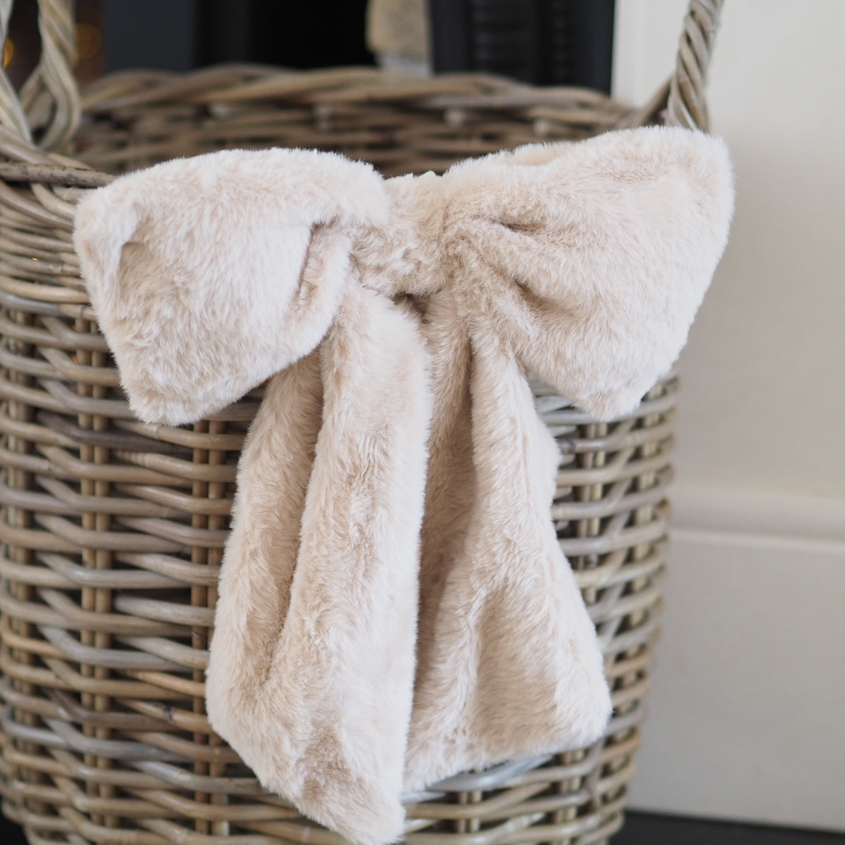 A soft, plush faux-fur bow in a light cream or oatmeal color, tied around a woven wicker basket.