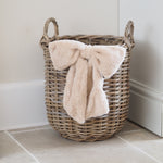 A natural wicker storage basket with two handles sits on a tiled floor against a white wall, holding a plush cream throw blanket tied in a large bow.