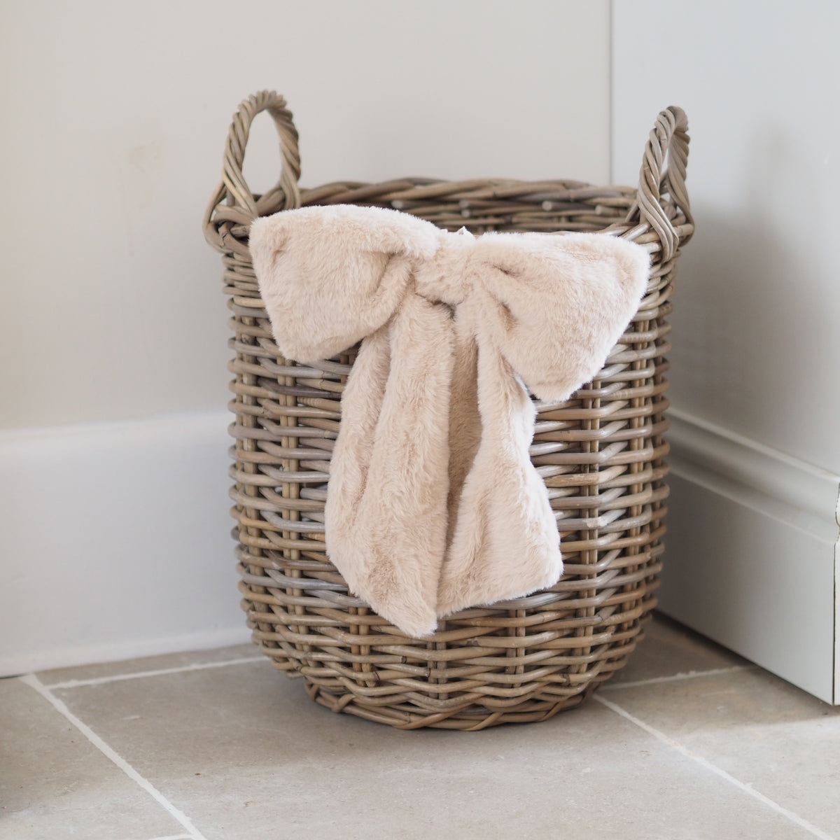 A natural wicker storage basket with two handles sits on a tiled floor against a white wall, holding a plush cream throw blanket tied in a large bow.