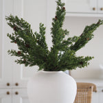 Green artificial pine branches with small brown pinecones arranged in a matte white ceramic vase, set against a softly blurred white kitchen background.