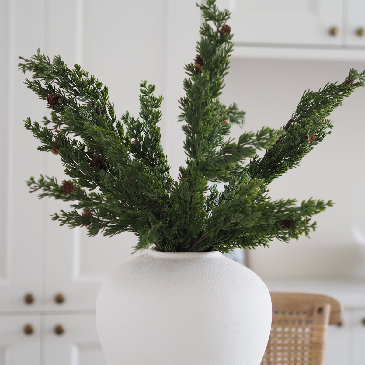 Green artificial pine branches with small brown pinecones arranged in a matte white ceramic vase, set against a softly blurred white kitchen background.
