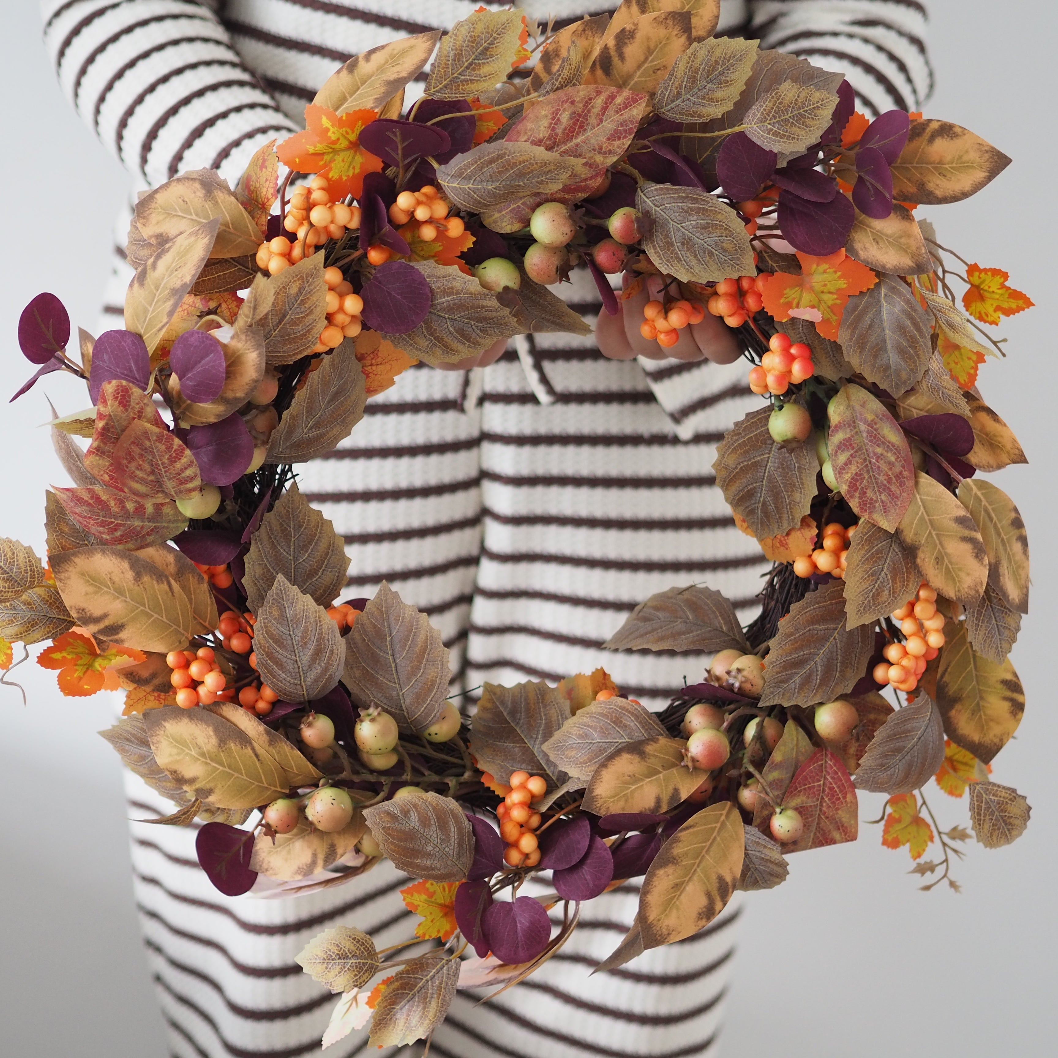 An autumn-themed artificial wreath with faux leaves in tan, brown, and purple, orange berries, and small pale green apples, held by a person wearing a black and white striped sweater against a light gray background.