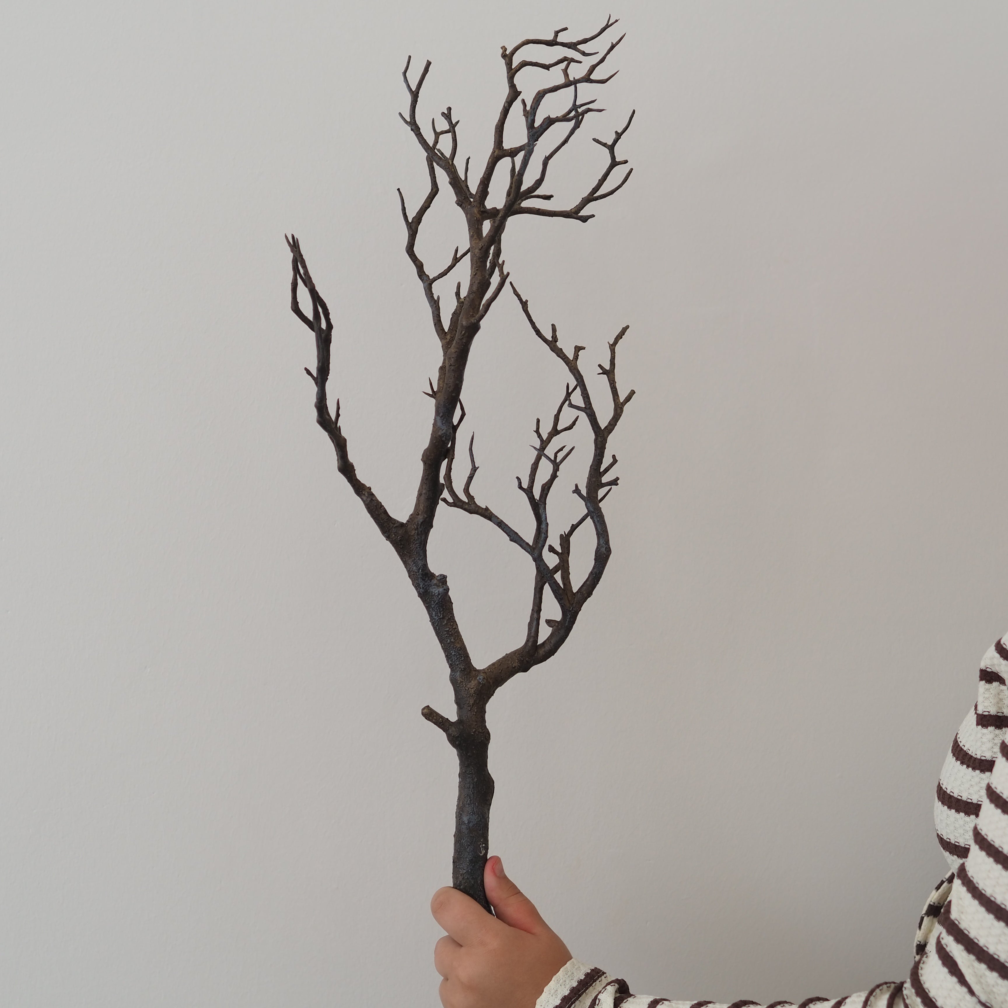A person holds a decorative artificial bare tree branch with dark brown and black textured branches against a plain light gray background.