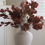 Artificial round-leaf foliage in muted burgundy and brown tones arranged in a matte white ceramic vase, set against a softly blurred indoor background.