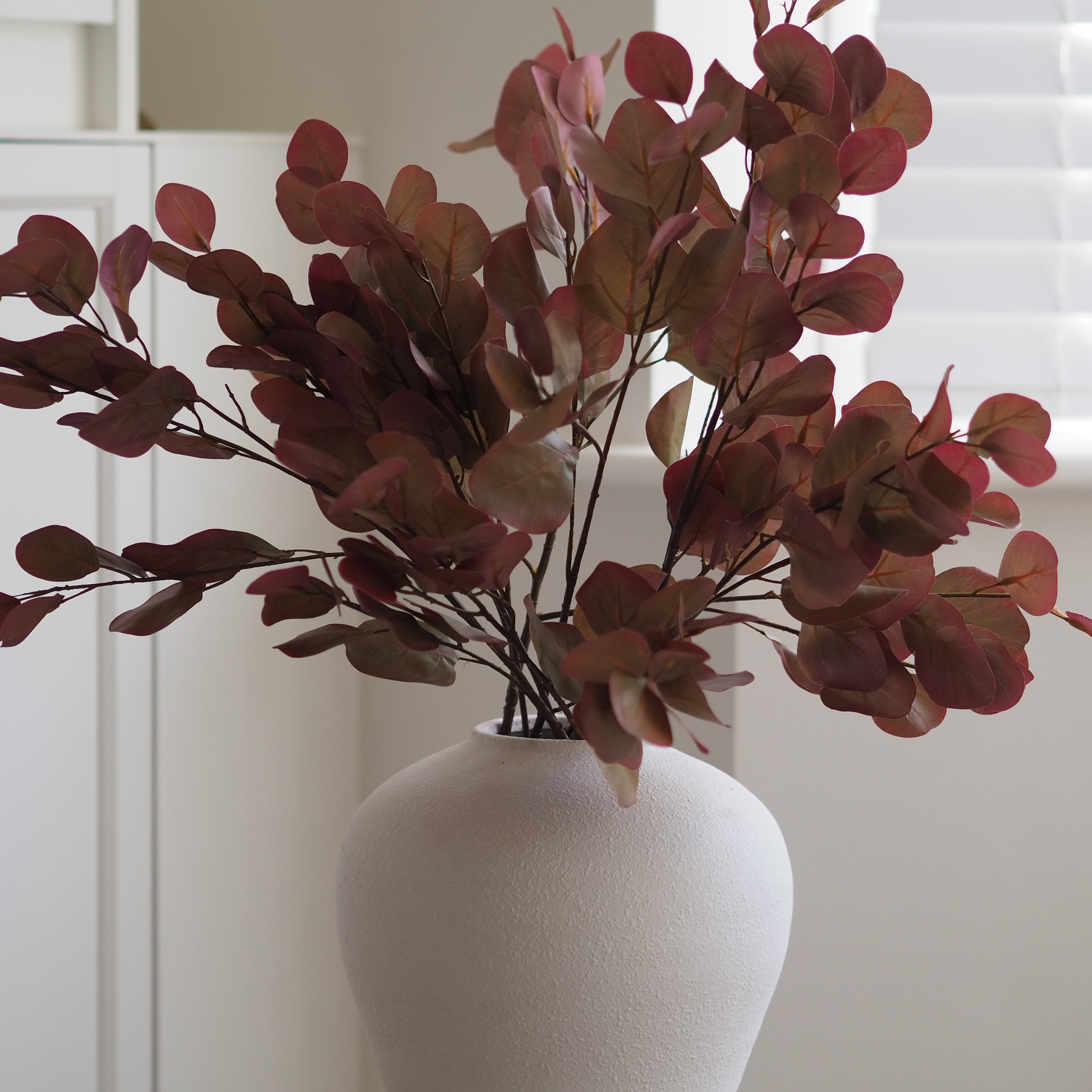 Artificial round-leaf foliage in muted burgundy and brown tones arranged in a matte white ceramic vase, set against a softly blurred indoor background.