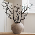 A decorative artificial tree branch arrangement with dark brown bare branches in a round textured beige ceramic pot, displayed on a light wood surface against a soft beige wall background.