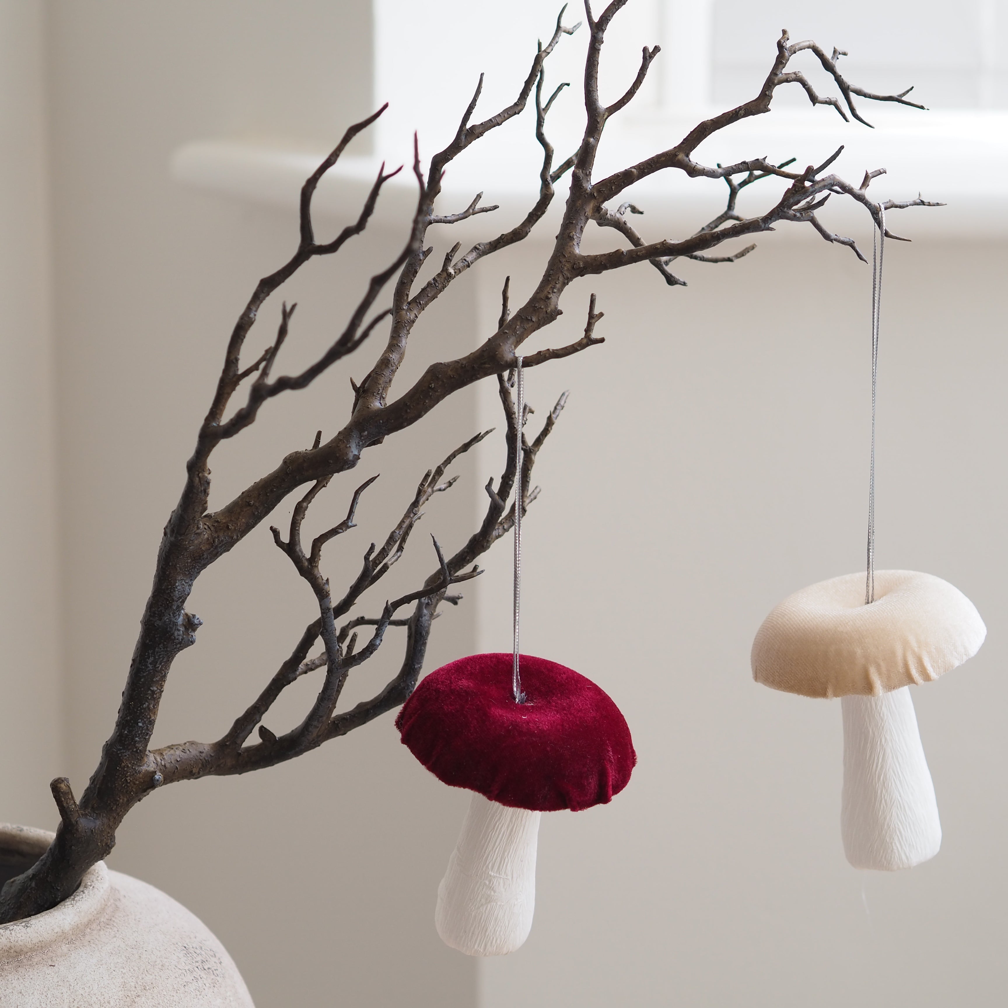 Two mushroom-shaped fabric ornaments in deep burgundy and cream, hanging from a decorative bare tree branch in a light-colored ceramic vase. Autumn decor Inspo 