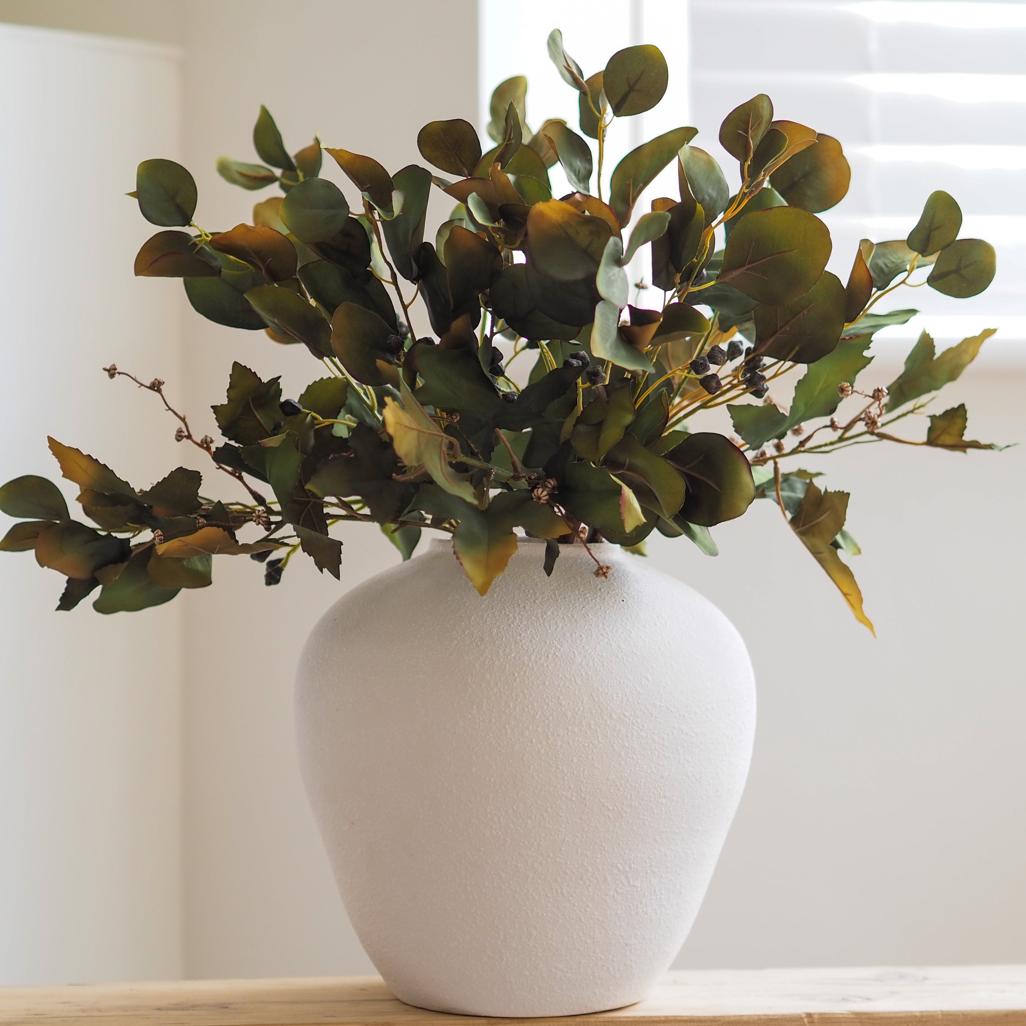 An artificial eucalyptus arrangement with green and brown leaves arranged in a textured white ceramic vase, set on a light wooden surface against a bright, softly lit background.