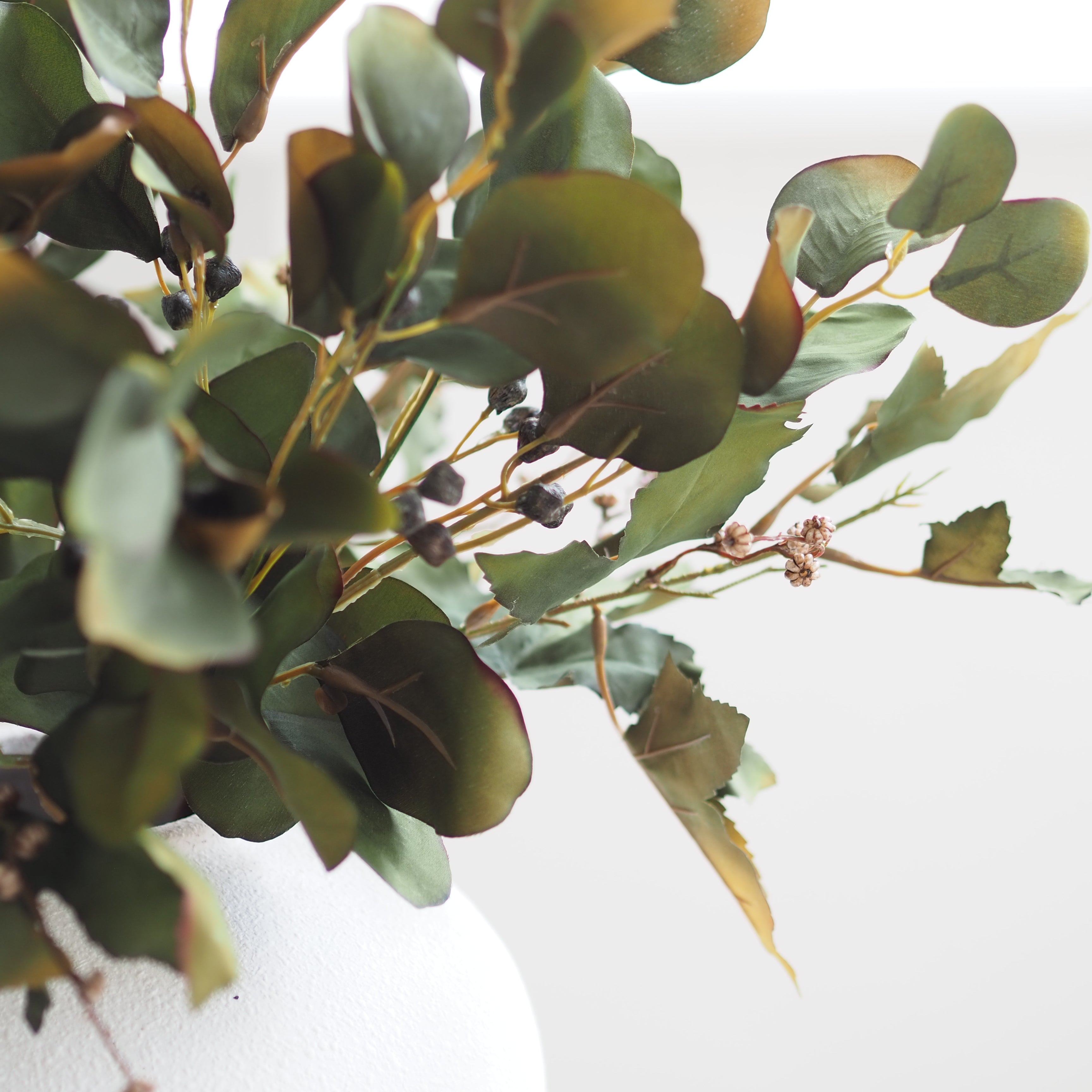 Artificial eucalyptus branch with round green leaves and small dark berries, arranged in a white textured vase against a bright white background.