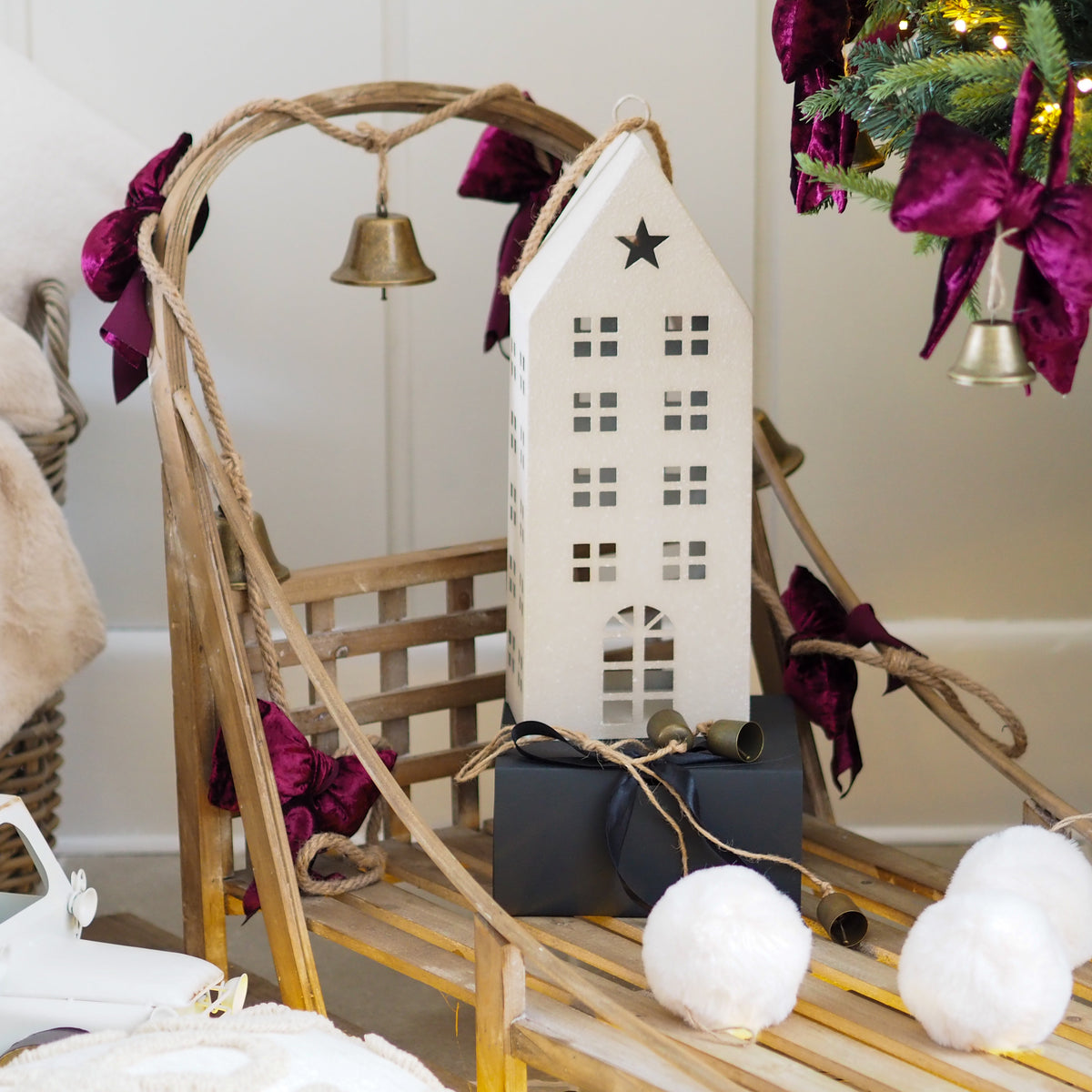 A white wooden house ornament with a star on the roof, displayed on a rustic wooden sled decorated with purple velvet bows and small brass bells, alongside a dark gift box and white pom-poms in a holiday setting.