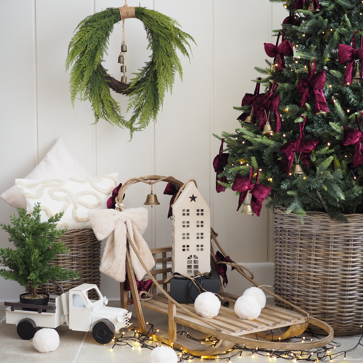 A styled Christmas scene with a green pine wreath hanging on a white paneled wall, a decorated Christmas tree in a wicker basket, a wooden sledding set with a small house, white pom-poms, a white truck, and warm string lights.