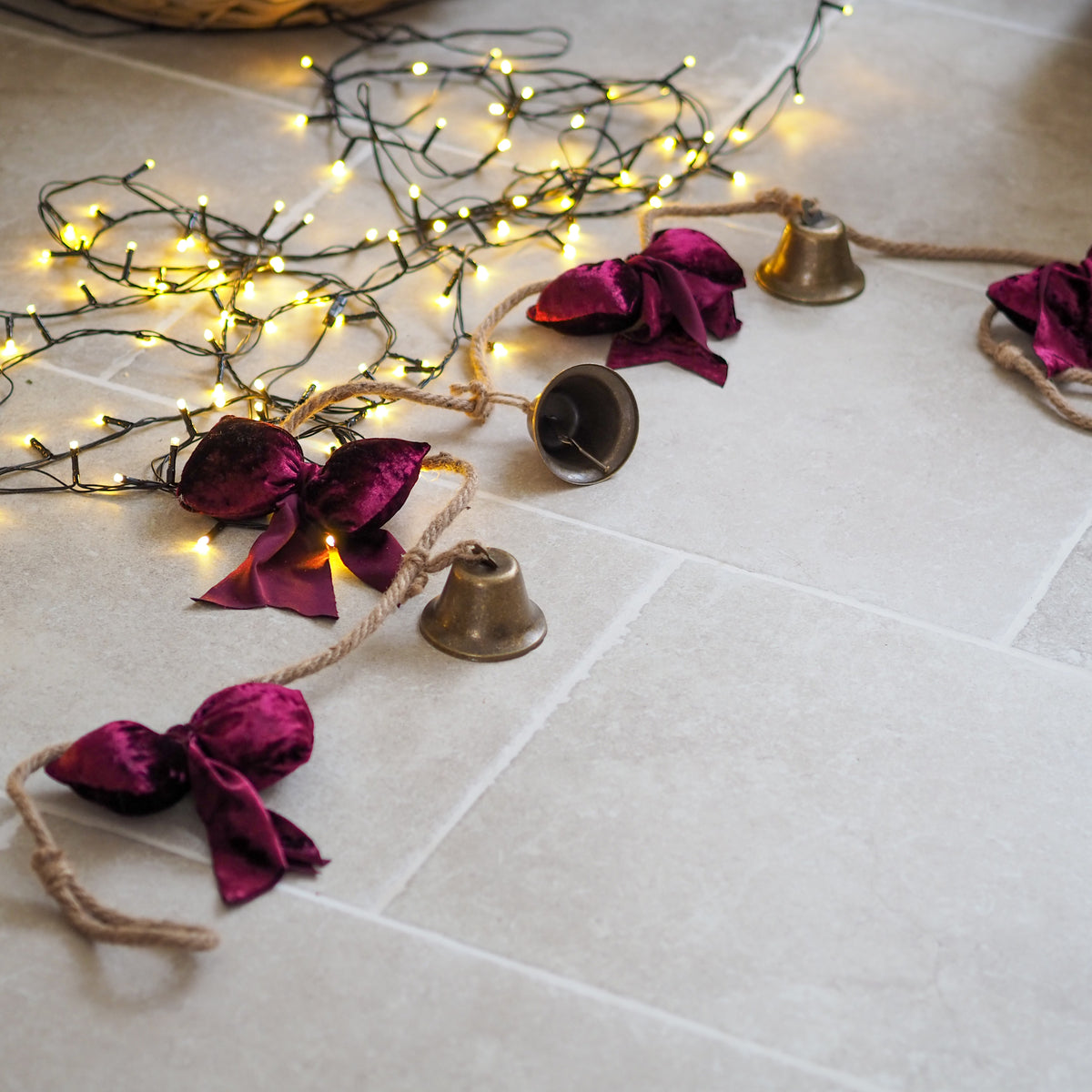 A festive garland draped across light beige floor tiles, featuring small brass bells, deep magenta velvet bows, and warm white string lights.