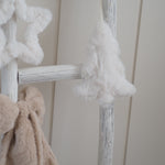 White faux fur Christmas tree ornament hanging on a distressed white wooden ladder next to a beige plush toy, against a cream paneled wall.