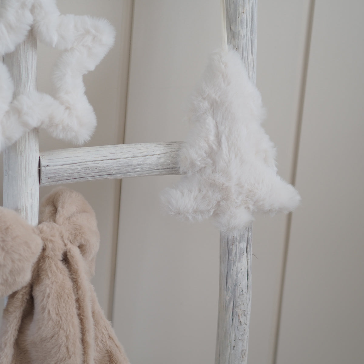 White faux fur Christmas tree ornament hanging on a distressed white wooden ladder next to a beige plush toy, against a cream paneled wall.