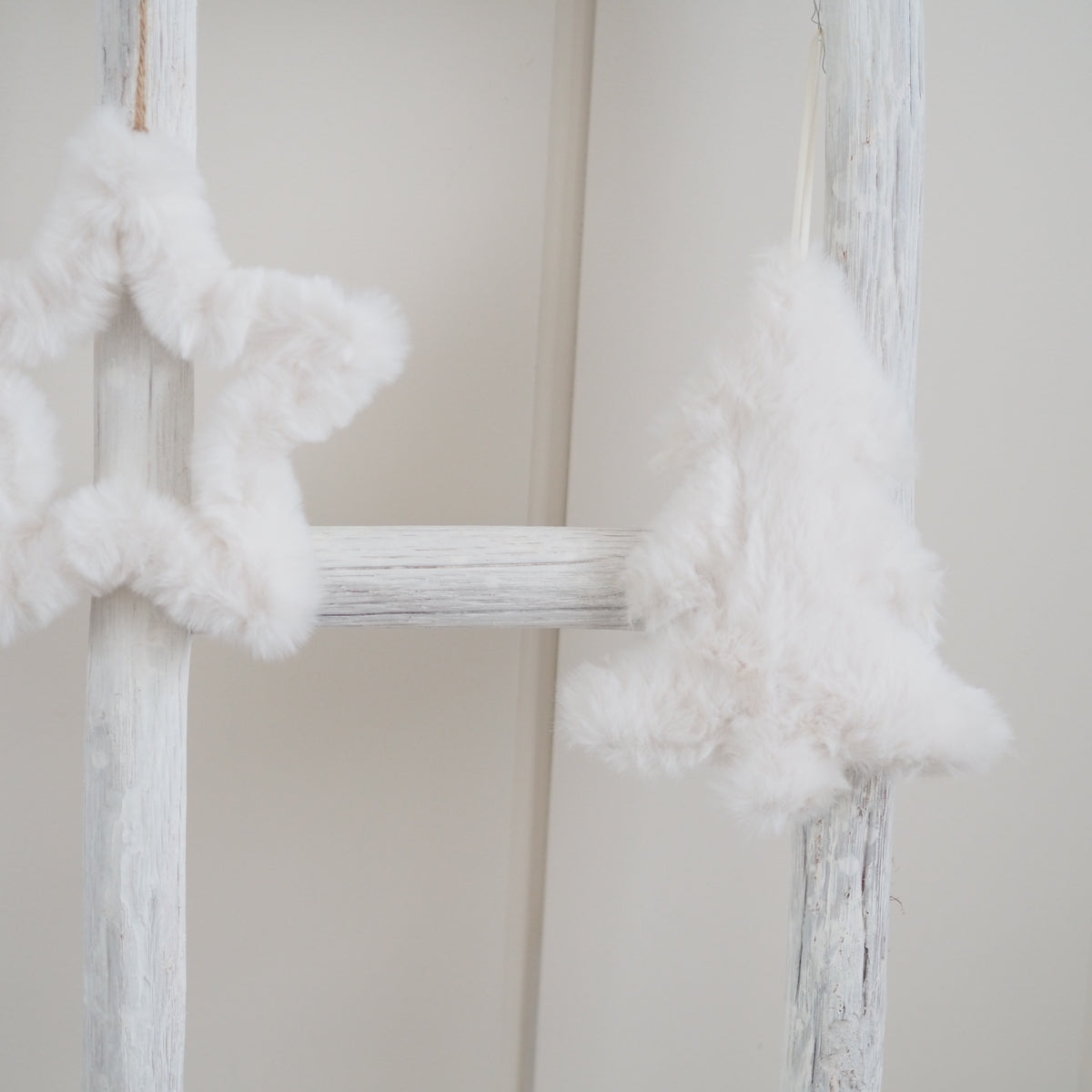 White fluffy faux-fur Christmas tree and star garlands hanging on a distressed white wooden ladder against a soft beige wall.