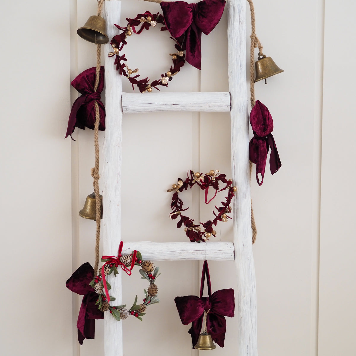 A rustic white wooden ladder decorated with three dark burgundy velvet wreaths, matching velvet bows, hanging gold bells on jute rope, and small pinecones, set against a cream paneled wall.