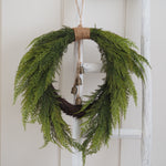 A green pine branch wreath hanging on a white wooden ladder, featuring a rustic jute-wrapped top and three small hanging brass bells.