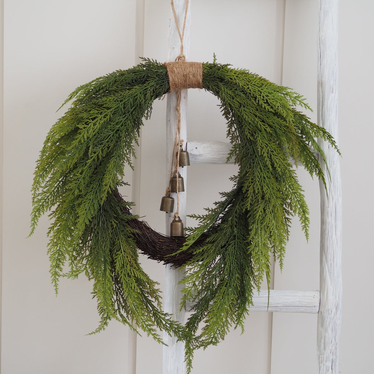 A green pine branch wreath hanging on a white wooden ladder, featuring a rustic jute-wrapped top and three small hanging brass bells.