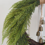 Green artificial cedar-style wreath with hanging bronze bells on a rustic white wooden post.