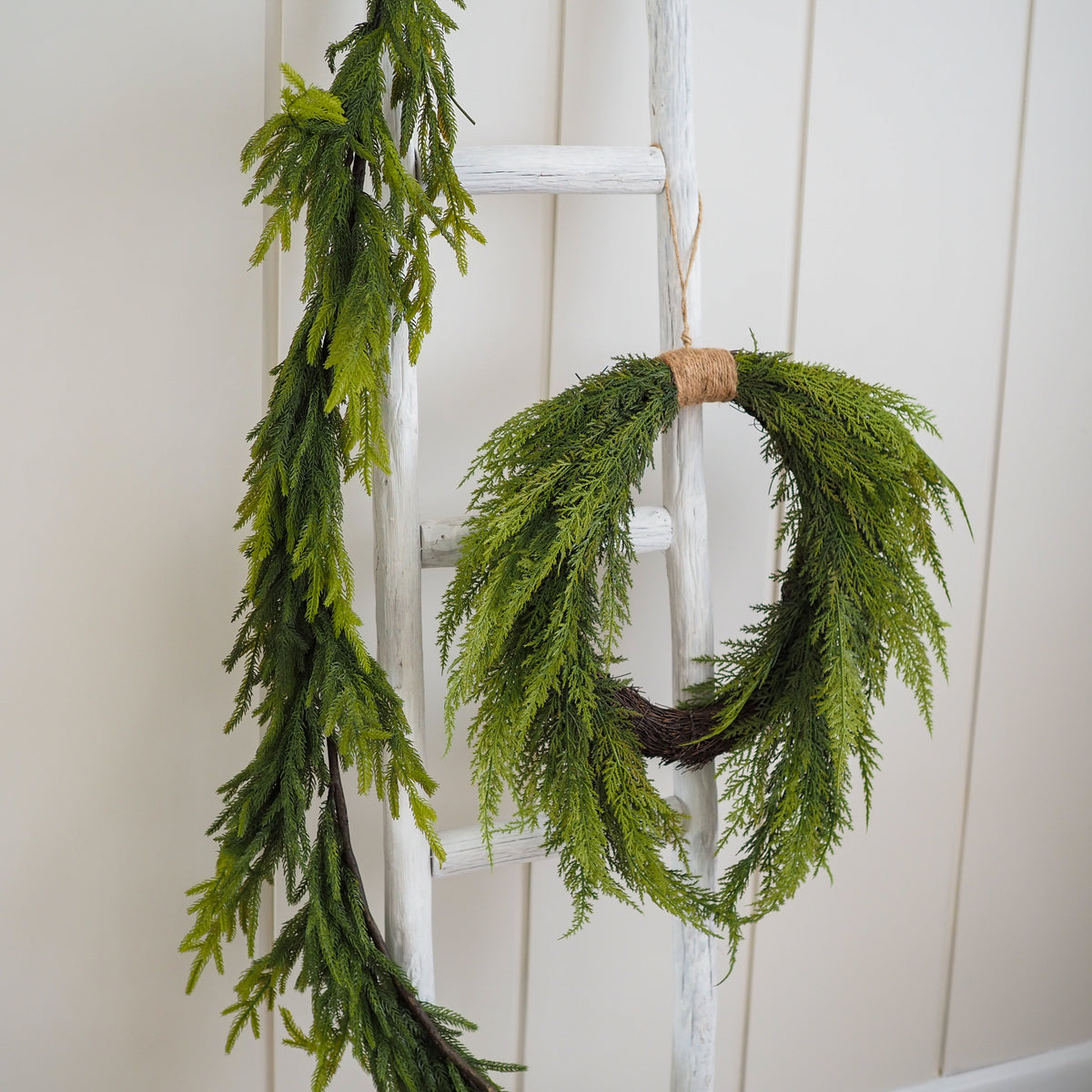 Green pine garland and matching hanging wreath displayed on a white wooden ladder against a light paneled wall.