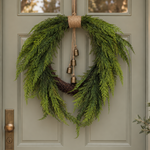A green evergreen wreath hanging on a gray paneled front door, with a natural jute rope top and four small hanging brass bells.