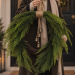 A person holds a large green evergreen wreath with hanging brass bells and a wrapped jute hanger, shown in a real doorway setting.